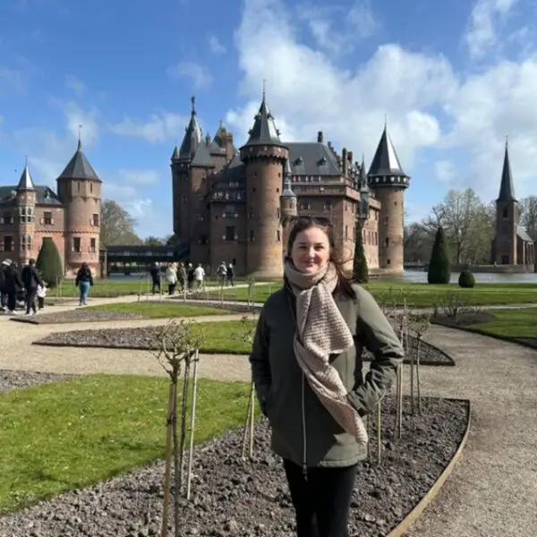 Persoon staat op een wandelpad voor een kasteel , met tuinen en bezoekers op de achtergrond.