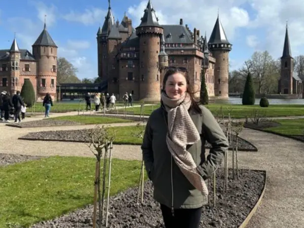 Persoon staat op een wandelpad voor een kasteel , met tuinen en bezoekers op de achtergrond.