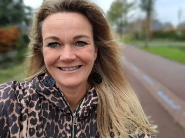 Persoon in een jas met dierenprint staat op een fietspad en kijkt richting de camera, met bomen en groen in de onscherpe achtergrond.
