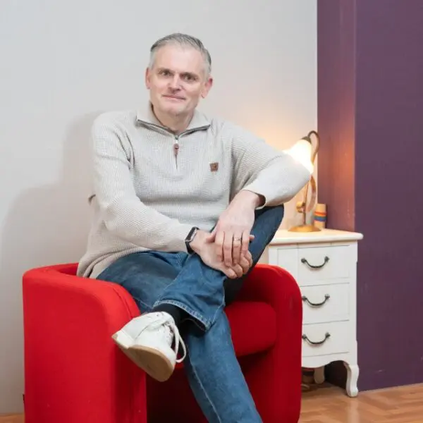 Persoon zit in een rode fauteuil in een woonruimte. Op de achtergrond staan een lamp en een ladekast.