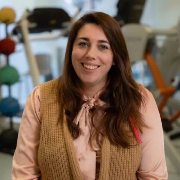 Portret van een persoon in een lichtroze blouse en beige vest, met op de achtergrond fitnessapparatuur.