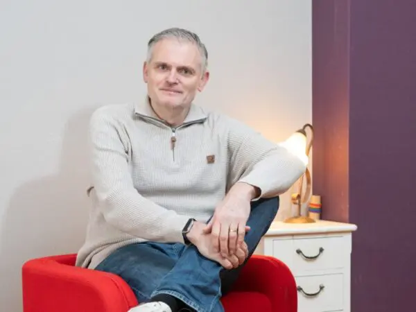 Persoon zit in een rode fauteuil in een woonruimte. Op de achtergrond staan een lamp en een ladekast.