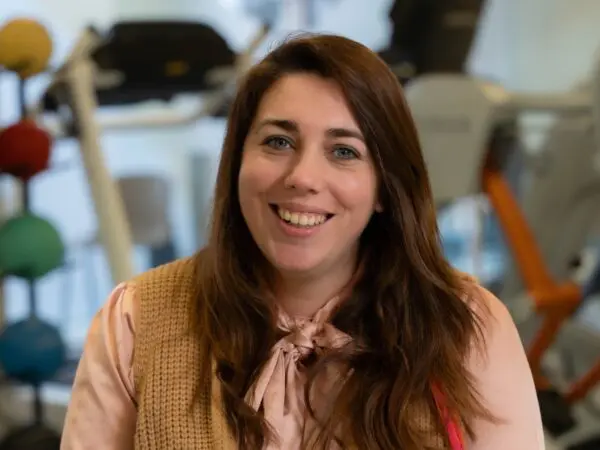 Portret van een persoon in een lichtroze blouse en beige vest, met op de achtergrond fitnessapparatuur.