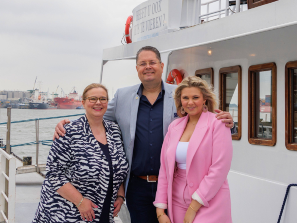 Drie personen staan op het dek van een schip in een haven. Op de achtergrond zijn schepen en haveninstallaties zichtbaar.