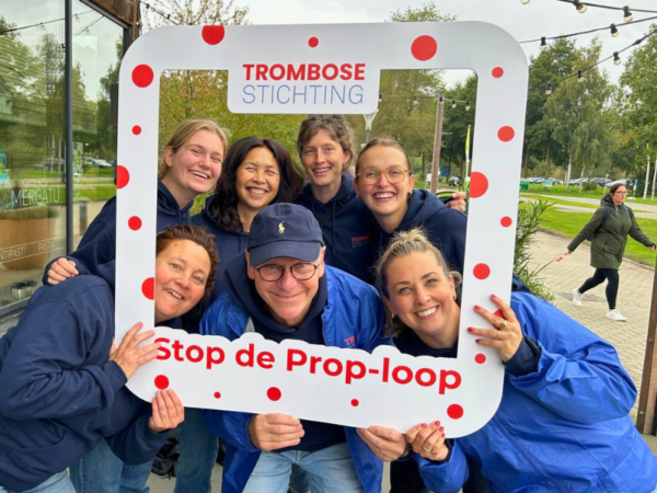 Groep van zeven personen poseert buiten achter een fotolijst van de Trombosestichting met de tekst 