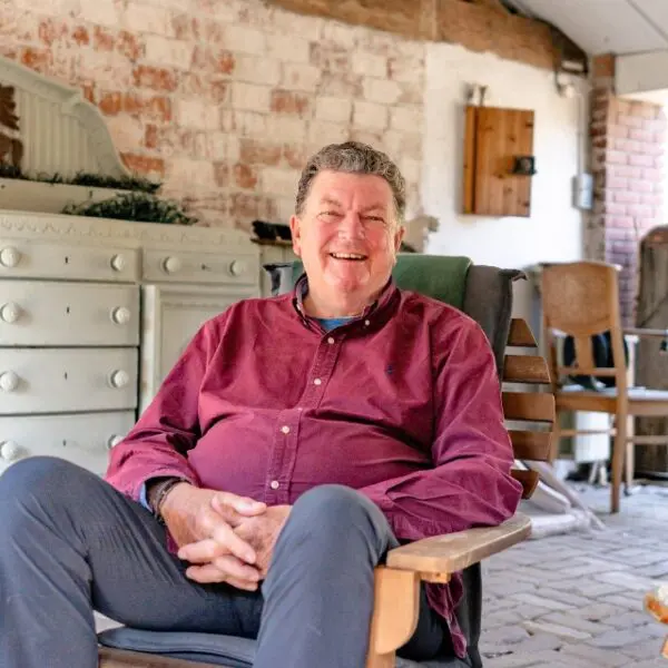 Persoon zit in een houten stoel op een overdekt terras met bakstenen muur en landelijk interieur op de achtergrond.
