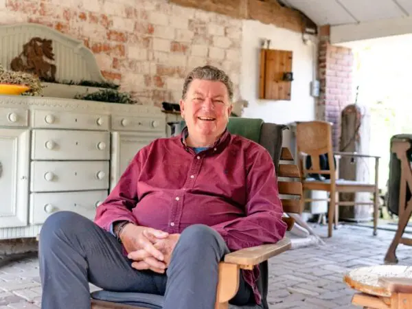 Persoon zit in een houten stoel op een overdekt terras met bakstenen muur en landelijk interieur op de achtergrond.
