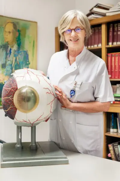 Persoon in een witte zorgjas staat naast een model van een oog in een kantoor met boekenkasten op de achtergrond.