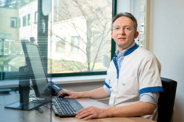 Persoon in een witte zorgjas zit achter een bureau met een computer in een kantoorruimte. Op de achtergrond zijn een raam en bomen zichtbaar.